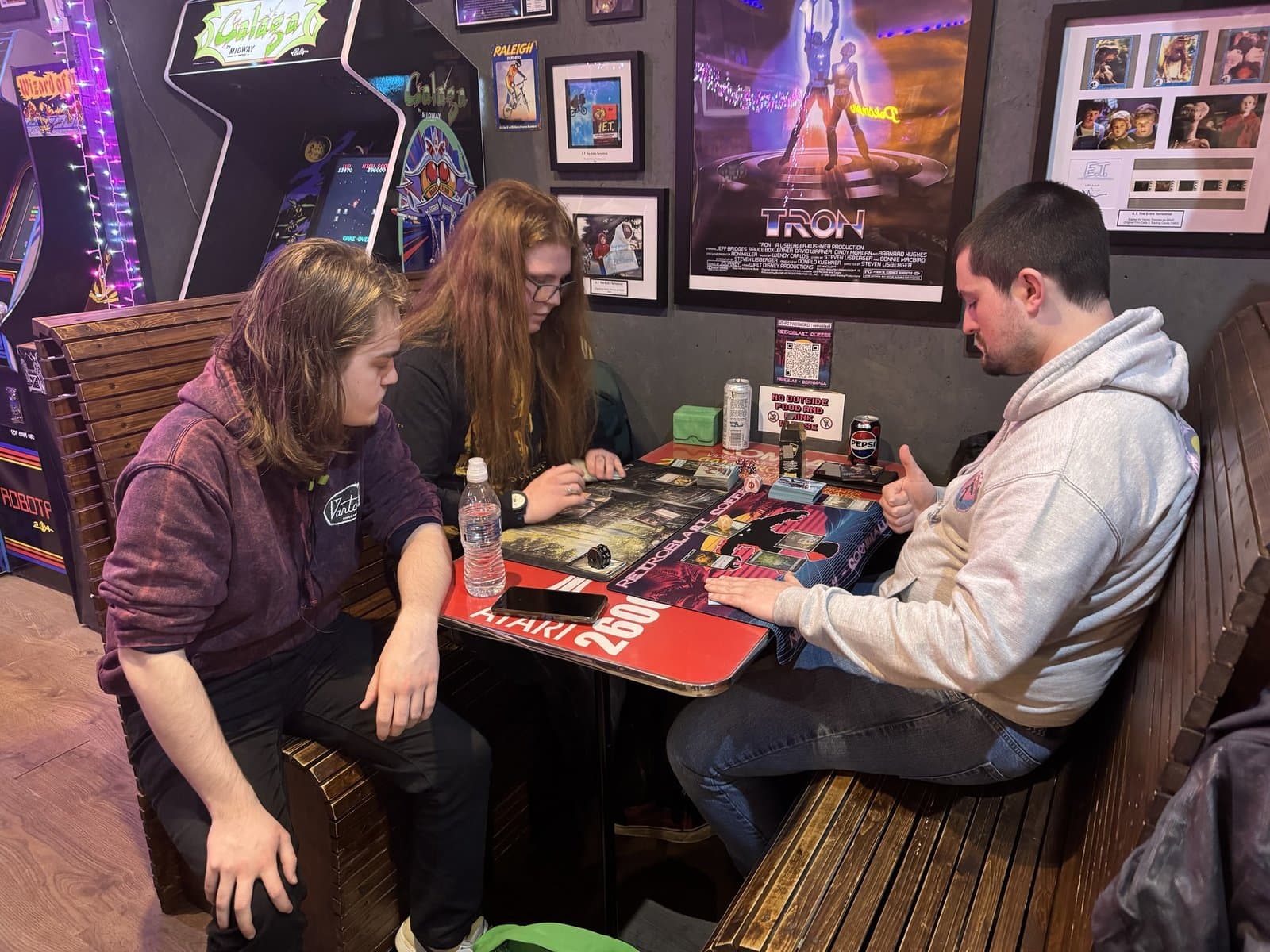 Board-game session under a Tron poster surrounded by arcade cabinets